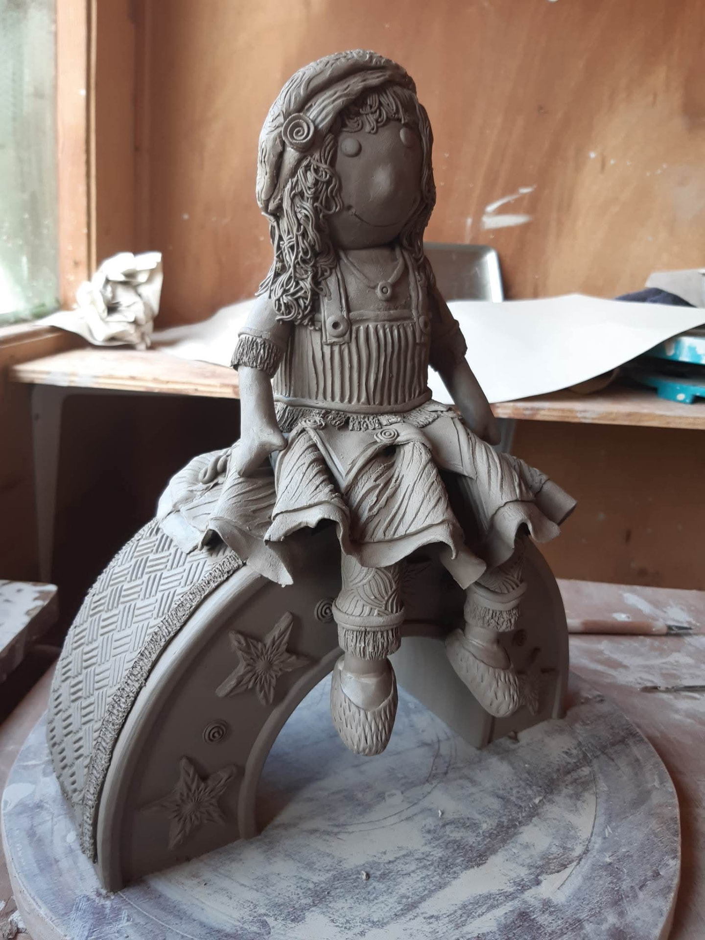 A clay girl figure seated on a carved crescent moon.