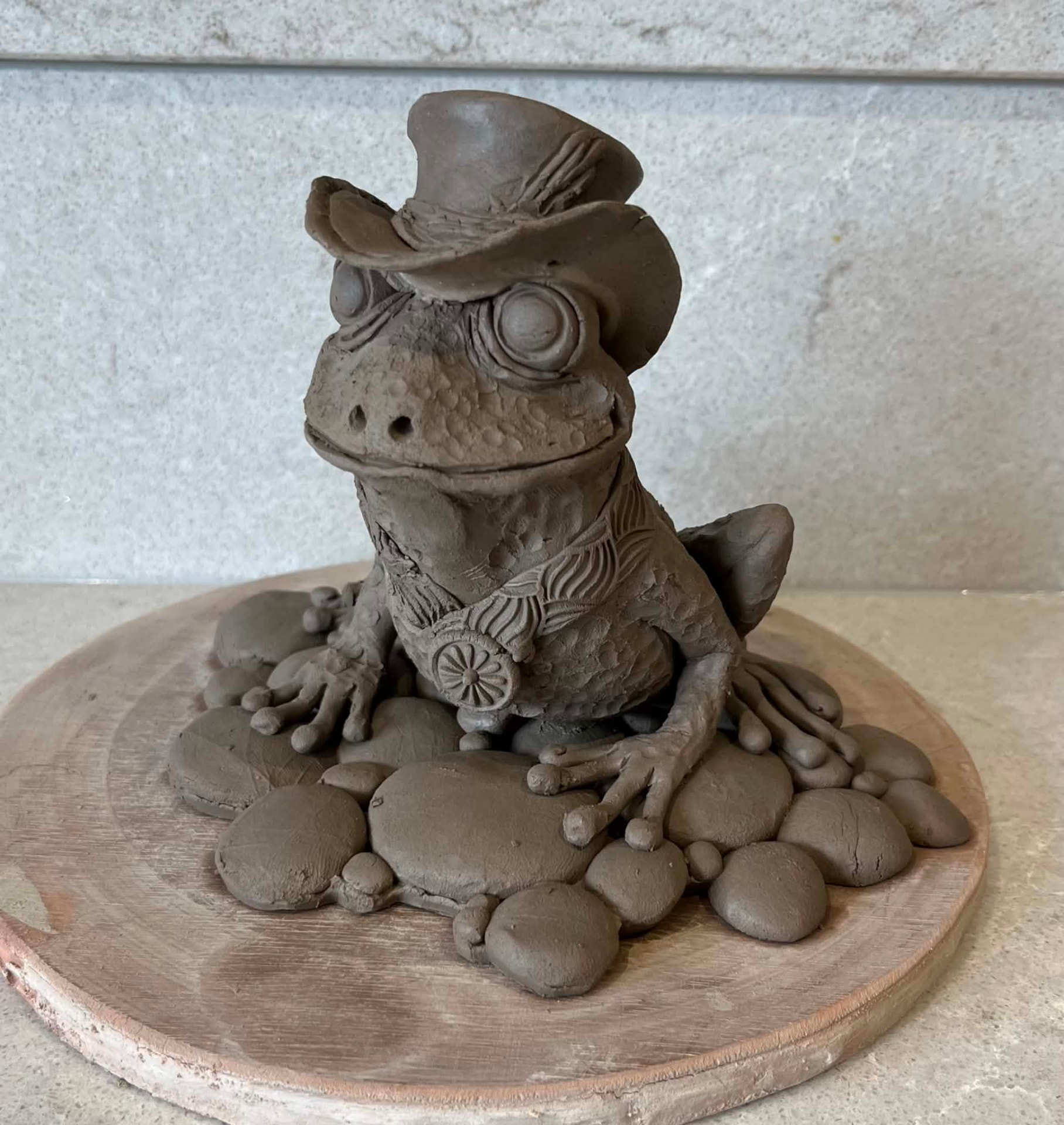 A dapper clay toad wearing a top hat and sitting on pebbles.